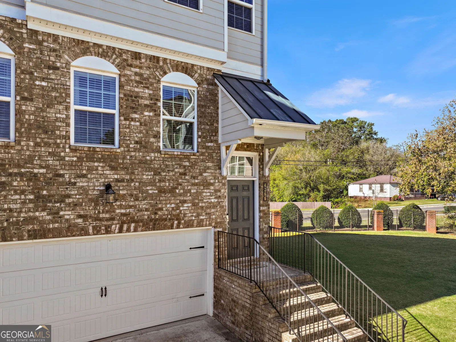 Front door and garage of the end unit, with the side lawn and a row of shrubs along the street